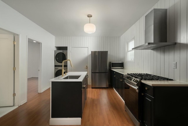 kitchen featuring wall chimney range hood, stacked washer and dryer, decorative light fixtures, a center island with sink, and appliances with stainless steel finishes