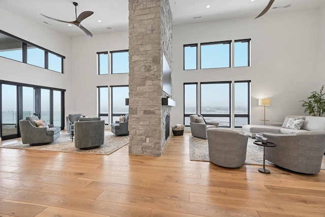 living room with a high ceiling, a water view, a stone fireplace, ceiling fan, and light hardwood / wood-style floors