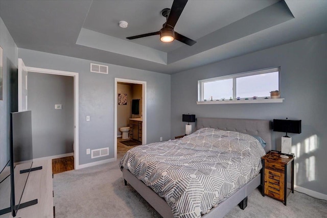 bedroom featuring ceiling fan, ensuite bathroom, light carpet, and a tray ceiling