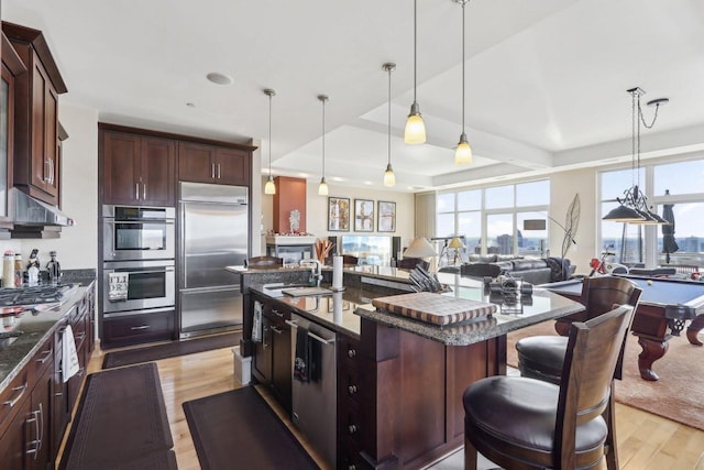 kitchen featuring light wood-type flooring, stainless steel appliances, a center island with sink, billiards, and hanging light fixtures