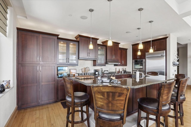 kitchen featuring stainless steel appliances, dark stone countertops, an island with sink, pendant lighting, and light wood-type flooring