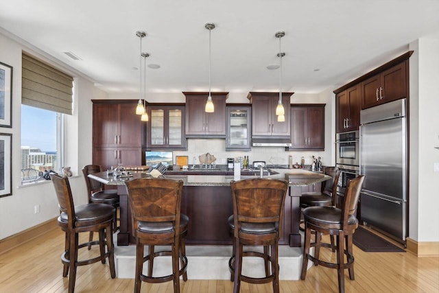 kitchen featuring a kitchen island with sink, dark stone counters, stainless steel built in fridge, decorative backsplash, and decorative light fixtures