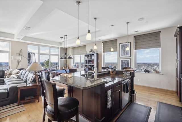 kitchen with dark stone countertops, decorative light fixtures, dark brown cabinets, a center island with sink, and light wood-type flooring