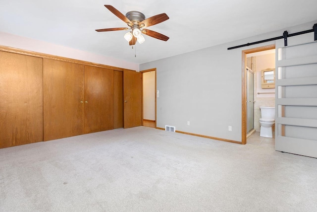 unfurnished bedroom featuring connected bathroom, ceiling fan, a barn door, light carpet, and a closet