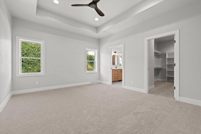 unfurnished bedroom featuring ceiling fan, ensuite bathroom, light colored carpet, a tray ceiling, and a walk in closet