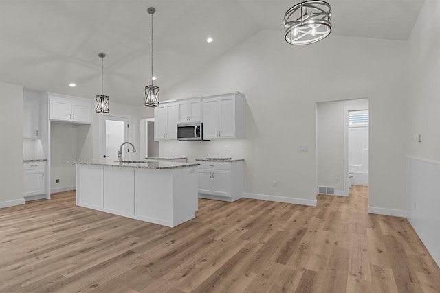 kitchen with light stone counters, high vaulted ceiling, pendant lighting, a kitchen island with sink, and white cabinets