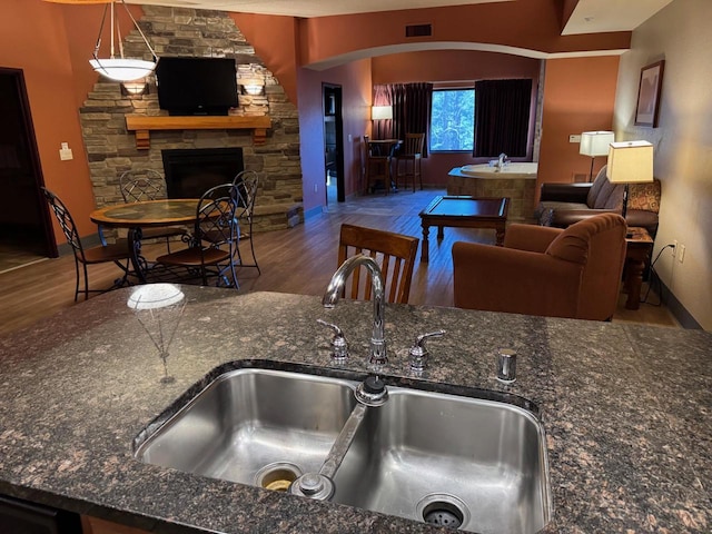 kitchen with sink, a stone fireplace, hardwood / wood-style floors, dark stone counters, and decorative light fixtures