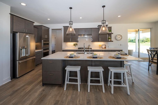 kitchen with decorative backsplash, dark hardwood / wood-style flooring, stainless steel appliances, a kitchen island with sink, and pendant lighting