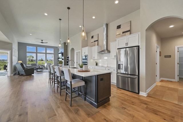 kitchen featuring white cabinetry, stainless steel appliances, wall chimney range hood, pendant lighting, and a center island with sink