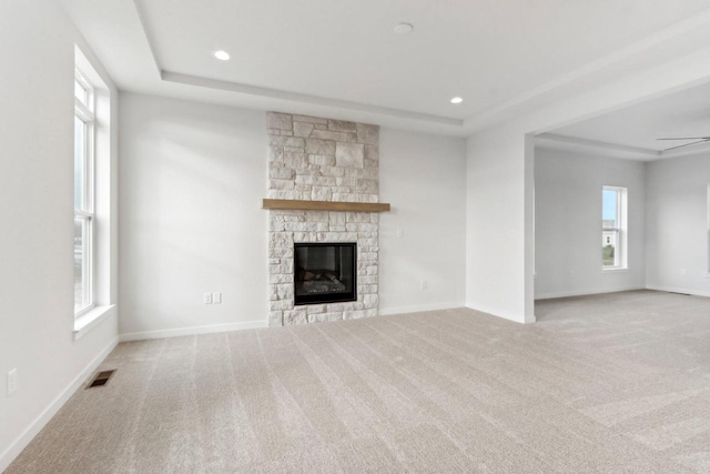 unfurnished living room with a raised ceiling, light colored carpet, a stone fireplace, and ceiling fan