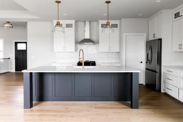 kitchen featuring stainless steel refrigerator with ice dispenser, an island with sink, decorative light fixtures, and wall chimney range hood