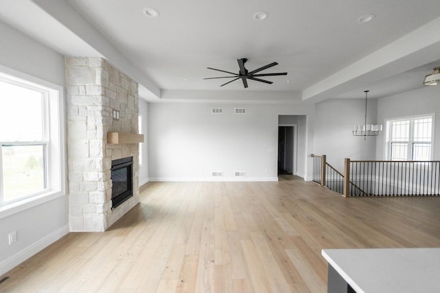 unfurnished living room with a stone fireplace, a healthy amount of sunlight, ceiling fan with notable chandelier, and light wood-type flooring