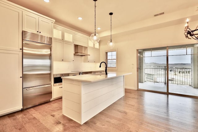 kitchen with wall chimney exhaust hood, stainless steel appliances, sink, pendant lighting, and an island with sink