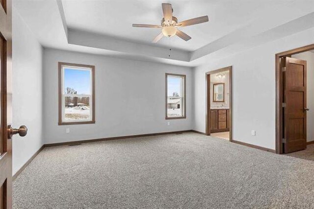 unfurnished bedroom featuring a raised ceiling, ensuite bath, ceiling fan, and multiple windows