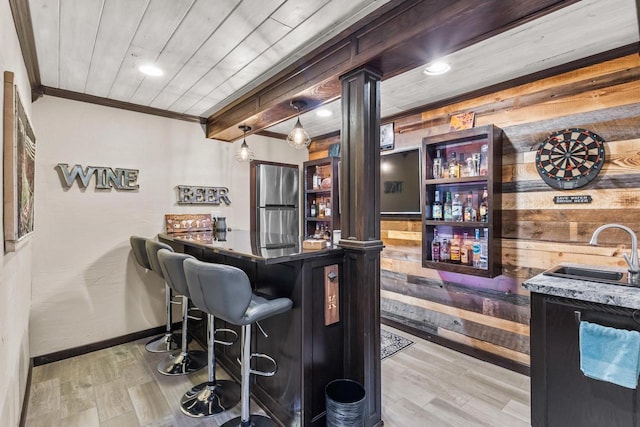 bar with crown molding, hanging light fixtures, sink, wooden walls, and stainless steel refrigerator