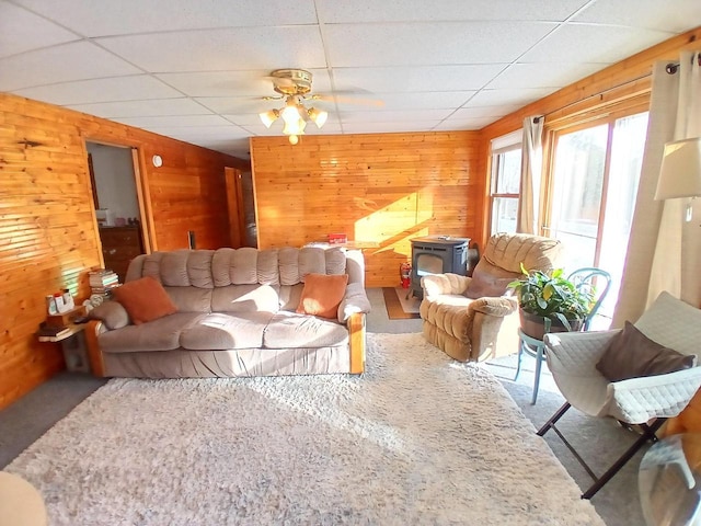 living room featuring a drop ceiling, a wood stove, ceiling fan, and wooden walls