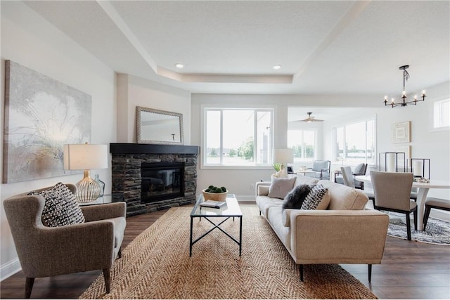 living room with a raised ceiling, a stone fireplace, dark hardwood / wood-style flooring, and ceiling fan with notable chandelier