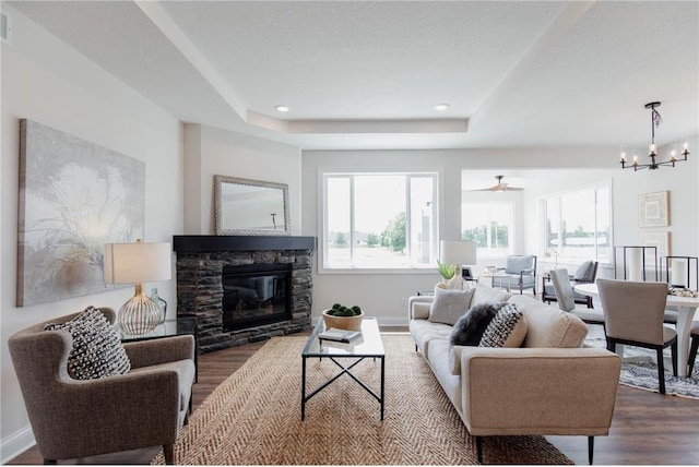 living room with a tray ceiling, a stone fireplace, dark wood-type flooring, and ceiling fan with notable chandelier