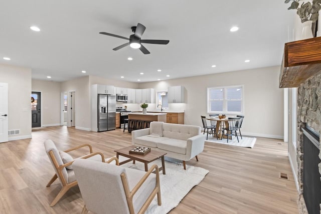 living room with light wood-type flooring, a stone fireplace, and ceiling fan