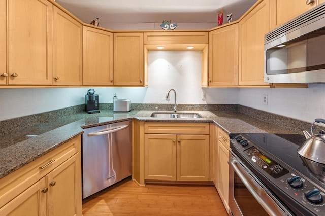 kitchen with appliances with stainless steel finishes, dark stone counters, sink, light brown cabinets, and light hardwood / wood-style flooring