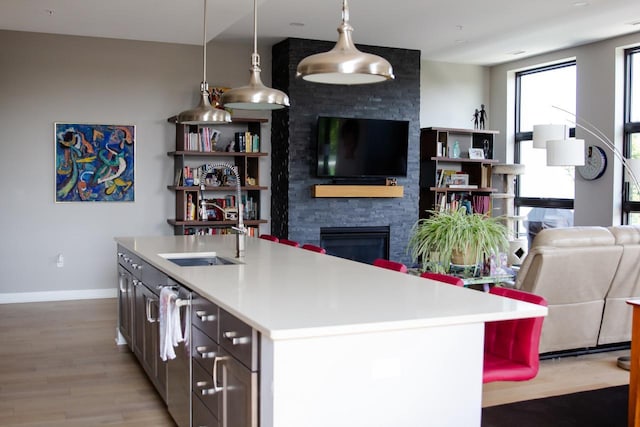 kitchen with light wood-type flooring, sink, pendant lighting, a stone fireplace, and an island with sink