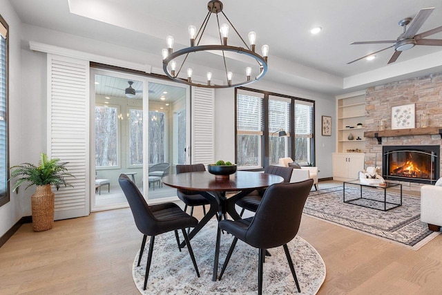 dining room with built in shelves, a raised ceiling, light hardwood / wood-style flooring, a fireplace, and ceiling fan with notable chandelier