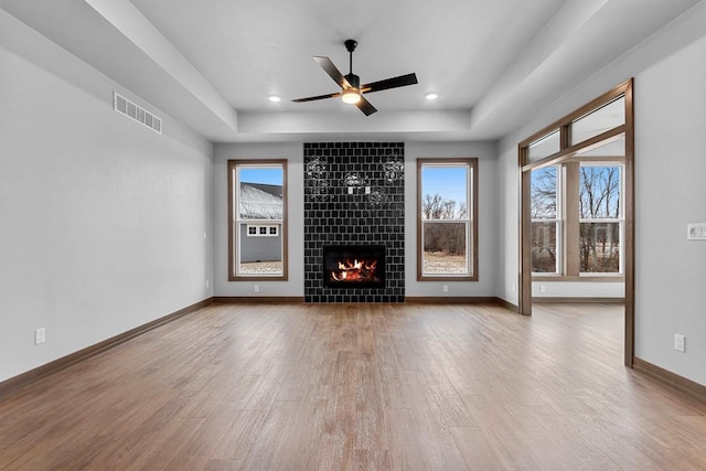 unfurnished living room featuring a tray ceiling, a tiled fireplace, ceiling fan, and wood-type flooring