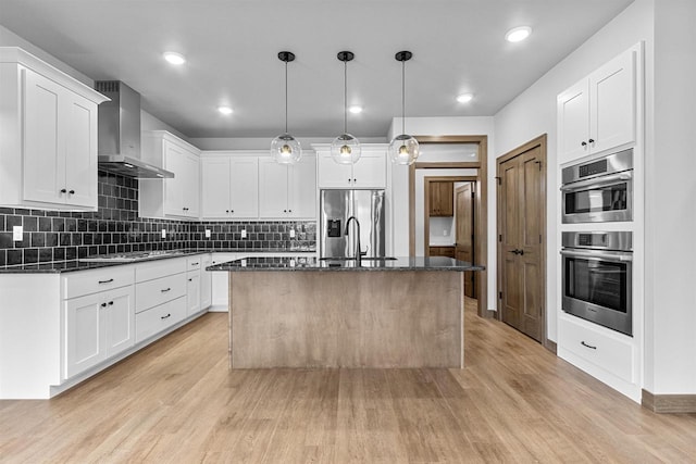 kitchen with white cabinets, wall chimney exhaust hood, an island with sink, decorative light fixtures, and light hardwood / wood-style floors