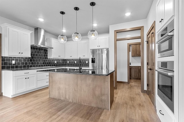 kitchen with white cabinets, stainless steel appliances, a center island with sink, and wall chimney range hood