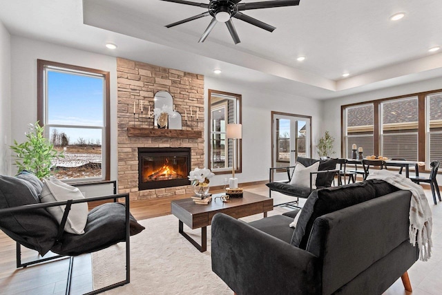 living room with ceiling fan, a stone fireplace, a raised ceiling, and light hardwood / wood-style flooring