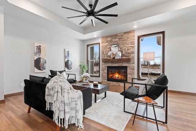 living room with a tray ceiling, a stone fireplace, ceiling fan, and light hardwood / wood-style floors