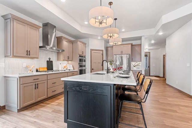 kitchen with wall chimney exhaust hood, light hardwood / wood-style flooring, an island with sink, pendant lighting, and appliances with stainless steel finishes