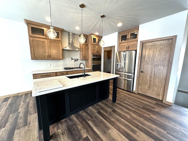 kitchen featuring wall chimney exhaust hood, stainless steel appliances, light stone counters, pendant lighting, and a center island with sink