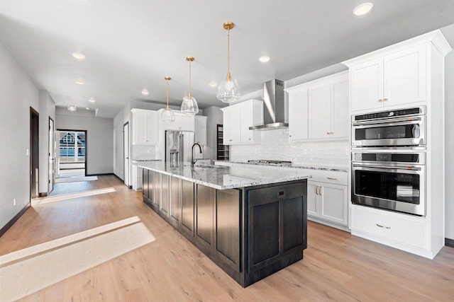 kitchen with wall chimney exhaust hood, hanging light fixtures, stainless steel appliances, a kitchen island with sink, and white cabinetry