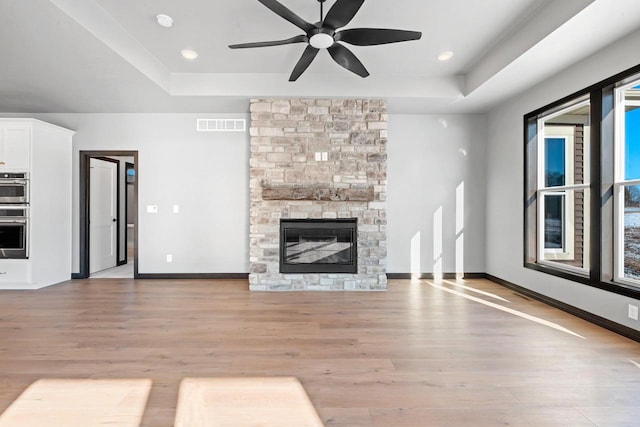 unfurnished living room featuring ceiling fan, light wood-type flooring, a stone fireplace, and a tray ceiling