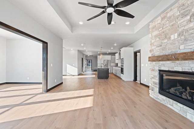 unfurnished living room featuring a stone fireplace, a raised ceiling, ceiling fan, sink, and light hardwood / wood-style flooring