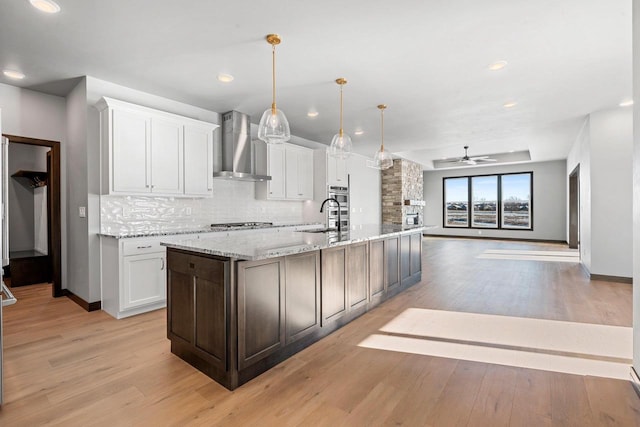 kitchen with a center island with sink, hanging light fixtures, ceiling fan, wall chimney exhaust hood, and white cabinets