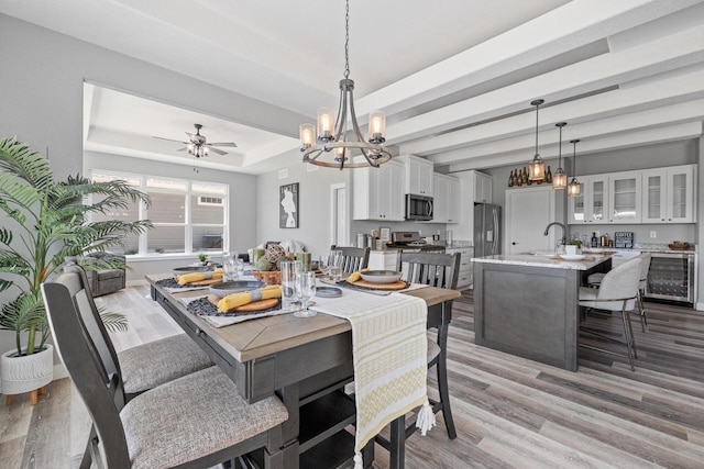 dining room featuring beverage cooler, ceiling fan with notable chandelier, baseboards, light wood-style floors, and a raised ceiling