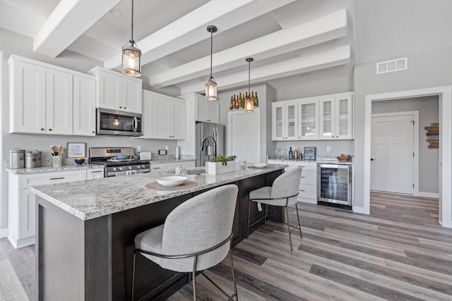 kitchen with beverage cooler, an island with sink, glass insert cabinets, stainless steel appliances, and white cabinetry