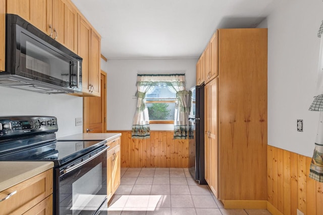 kitchen with black appliances, light tile patterned flooring, light brown cabinetry, and wood walls