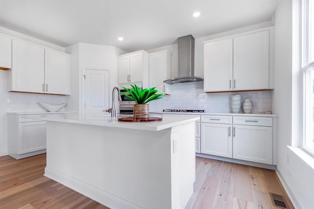 kitchen featuring white cabinetry, light wood-type flooring, decorative backsplash, a center island with sink, and wall chimney range hood