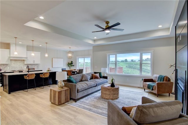 living room with ceiling fan with notable chandelier, a tray ceiling, and light hardwood / wood-style flooring