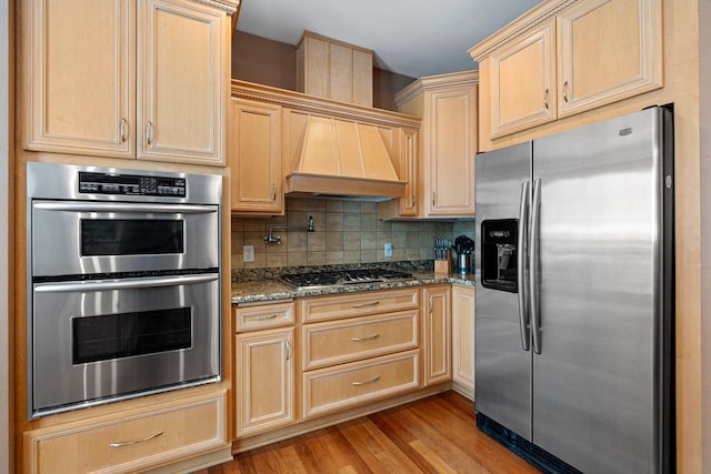 kitchen with stainless steel appliances, decorative backsplash, light brown cabinets, light wood-type flooring, and dark stone counters