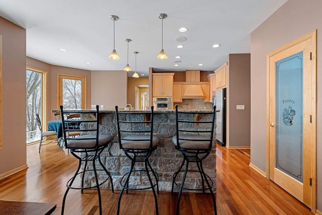 kitchen with tasteful backsplash, decorative light fixtures, stainless steel appliances, custom range hood, and light brown cabinetry