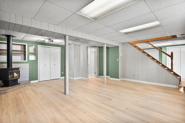 basement with a wood stove, a paneled ceiling, and light hardwood / wood-style floors