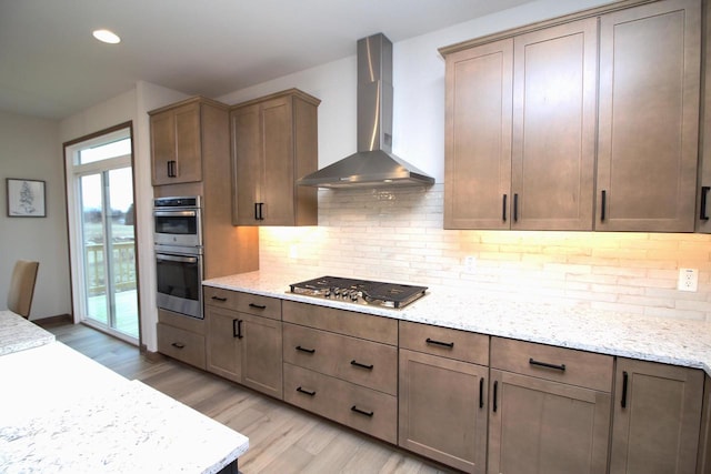 kitchen with decorative backsplash, light hardwood / wood-style flooring, stainless steel appliances, wall chimney exhaust hood, and light stone counters