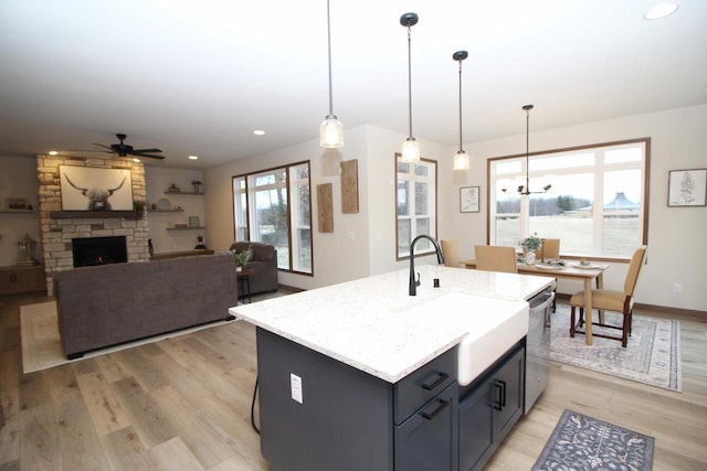 kitchen with ceiling fan, a fireplace, a center island with sink, stainless steel dishwasher, and hanging light fixtures