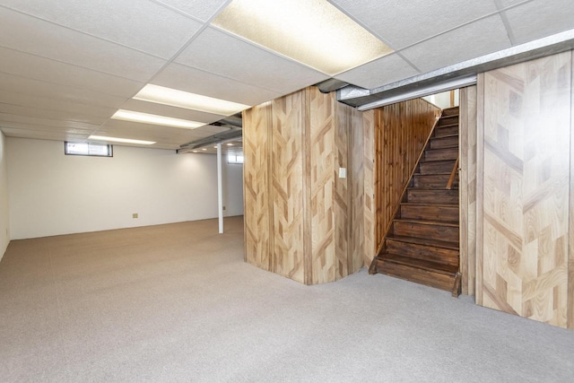 basement with a drop ceiling, wood walls, a healthy amount of sunlight, and carpet flooring