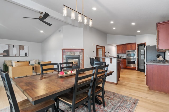 dining space with ceiling fan, a fireplace, light hardwood / wood-style floors, beam ceiling, and high vaulted ceiling