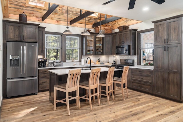 kitchen with stainless steel appliances, a kitchen island with sink, light wood-type flooring, dark brown cabinets, and beamed ceiling